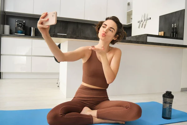 Beautiful girl in yoga sportswear, sits on her floor at home, takes selfie on yoga mat, recording workout vlog.