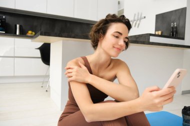 Spor ve refah. Spor salonundaki kadın portresi, evdeki beden eğitimi hocası yoga minderinde oturur ve gülümseyen yüzüyle akıllı telefonuna bakar..