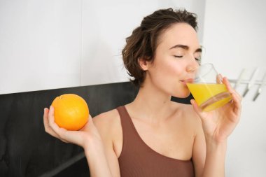 Sağlıklı yaşam tarzı ve spor. Güzel, gülümseyen bir kadın, taze portakal suyu içiyor ve elinde meyve tutuyor, evde egzersiz yaptıktan sonra vitamin içeceğinin tadını çıkarıyor, mutfakta duruyor..
