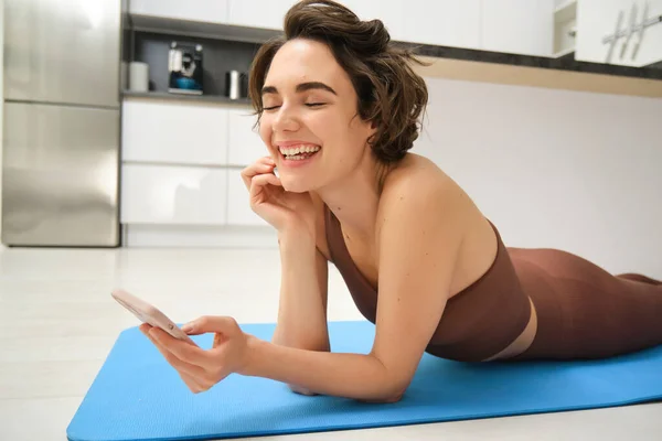 Close up portrait of fitness girl, lying on yoga mat and laughing, using smartphone, records workout exercises on mobile phone, does training at home.