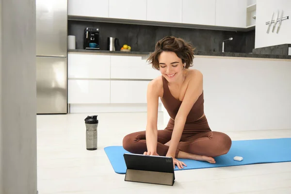 Young fit and healthy woman, yoga instructor, fitness girl in tracksuit, sits on floor rubber mat, using digital tablet, watches video pilates tutorial, stretching exercises.
