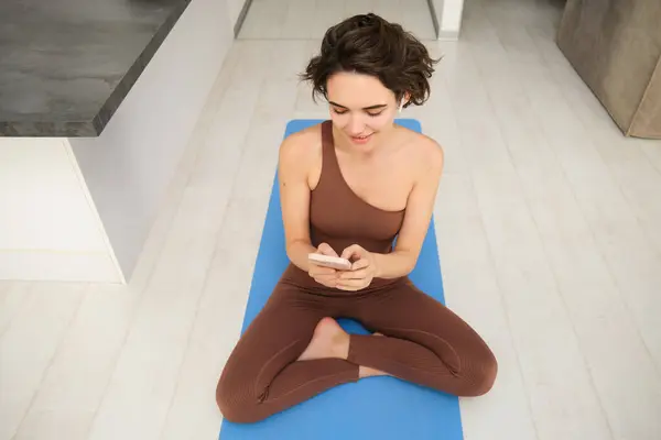 Portrait of fitness girl listens music, workout from home on yoga mat, uses smartphone sport app, watches training session, online exercises.