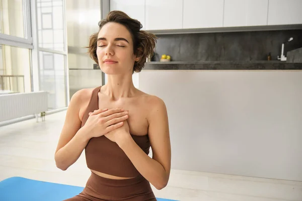 Sukhasana Hands To Heart. Portrait of young brunette fitness woman, girl holds hans on chest, doing yoga at home in sport leggins and bra, sitting on floor indoors.