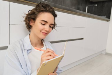 Günlüğe yazan güzel genç bir kadının portresi, planlamacıya notlar eklemek, yere oturup düşünmek, günlüğünü okumak, mutlu bir yüz ifadesiyle gülümsemek..