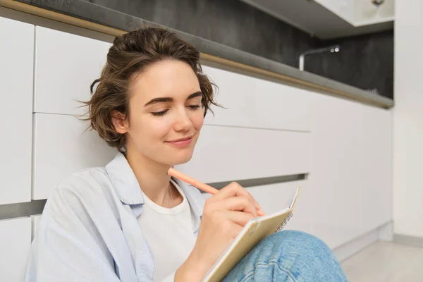Close up portrait of young woman writing in her journal, feeling creative, sitting with planner, notebook and pen on floor, studying, doing her homework at home.