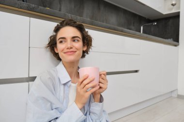 Şefkat ve rahatlama. Genç bir kadın kahve içiyor ve gülümsüyor, bir fincan çayla rahatlıyor, mutfakta yerde oturuyor..