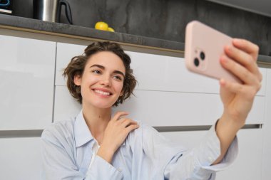 Genç bir kadının portresi mutfak zemininde telefonuyla oturuyor, akıllı telefonuyla fotoğraf çekiyor uygulama filtreleriyle, cep telefonuyla poz veriyor..