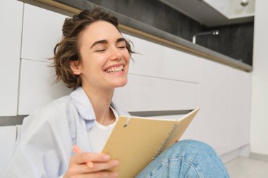 Günlüğüne yazan, yaratıcı hisseden, yerde planlayıcı, defter ve kalemle oturan, ders çalışan, ödevlerini evde yapan genç bir kadının portresini kapat..