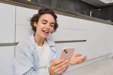 Mutlu genç bir kadının portresi, yepyeni mutfağını gösteriyor, yere oturuyor, mobilyaları gösteriyor ve gülümsüyor, akıllı telefondaki video sohbetleri. Teknoloji ve yaşam tarzı kavramı