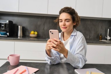 Güzel gülümseyen bir kadının portresi akıllı telefonuyla evde oturuyor, cep telefonu uygulaması kullanıyor, erzak siparişi veriyor..