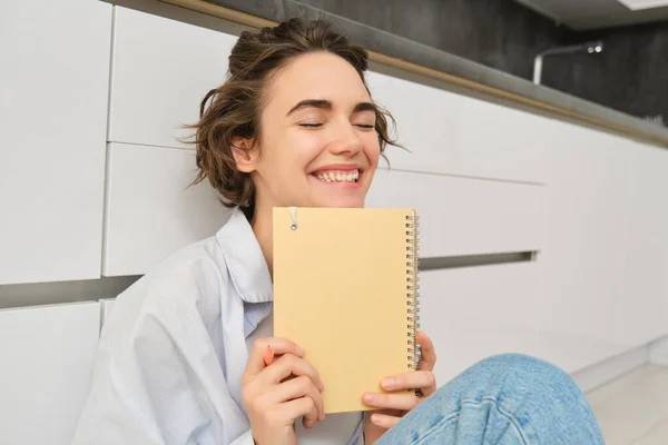 Portrait of happy, carefree woman, writes in her journal and laughs, holds her diary, reads something in notebook, sits on floor indoors. Lifestyle and leisure concept