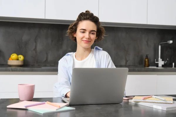 stock image Productive young woman works from home, looks at her laptop, sits in her kitchen with papers and cup of coffee.