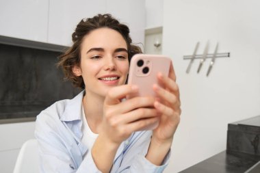 Akıllı telefonuna bakan, mesaj okuyan, evde oturup internetten alışveriş yapan, mobil uygulamalardaki hizmetleri ödeyen genç bir kadının görüntüsü.