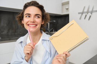 Genç yaratıcı kadının portresi, defterini tutuyor ve gülümsüyor, günlüğüne notlar yazıyor, ajandasına yazıyor, mutfakta oturuyor ve gülümsüyor..