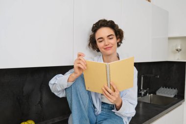 Yaşam tarzı ve insan kavramı. Genç bir kadın hafta sonunun keyfini çıkarıyor, günlüğünü okuyor, mutfak tezgahına oturuyor ve gülümsüyor..