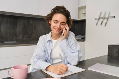 Evden çalışan bir kadının görüntüsü, bir telefon görüşmesi sırasında bilgi yazmak, birini aramak, akıllı telefon kullanmak..