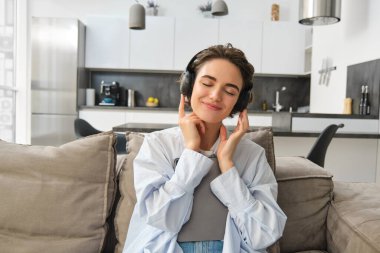 Güzel, tasasız bir kızın portresi kulaklıkla müzik dinliyor, neşeli bir gülümsemeyle tablet tutuyor, evdeki kanepede oturuyor. Boşluğu kopyala