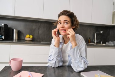 Kadın portresi cep telefonuyla konuşurken, mutfakta akıllı telefonuyla otururken ve kafası karışmış bir kalemi ısırırken..