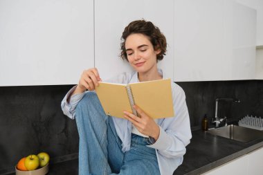 Genç modern kadın portresi mutfakta oturuyor, sayfaları çeviriyor, günlük kitabı okuyor, mutlu bir şekilde gülümsüyor. İnsanlar ve yaşam tarzı kavramı