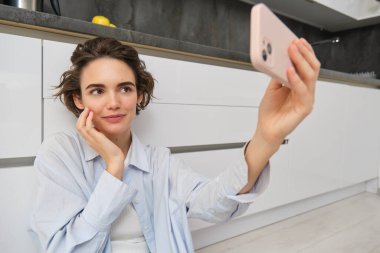Genç bir kadının portresi mutfak zemininde telefonuyla oturuyor, akıllı telefonuyla fotoğraf çekiyor uygulama filtreleriyle, cep telefonuyla poz veriyor..