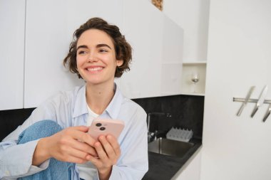 Güzel bir kızın hayat tarzı fotoğrafı, akıllı telefonuyla evde oturuyor ve gülümsüyor, yüzünde mutlu bir duyguyla başka yere bakıyor..