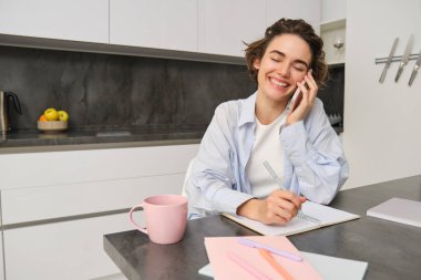 Samimi, gülümseyen genç bir kadının portresi, cep telefonuyla konuşmak, akıllı telefondaki konuşmalar sırasında gülmek, not almak, deftere not almak, mutfakta oturmak..