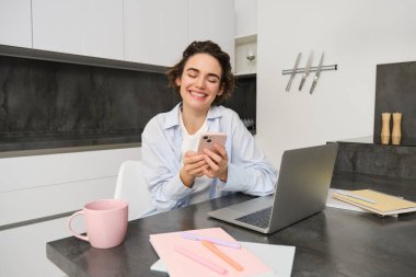 Evde akıllı telefonunu kullanan genç bir kadının görüntüsü. Kız cep telefonuyla mutfakta oturuyor ve gülümsüyor..