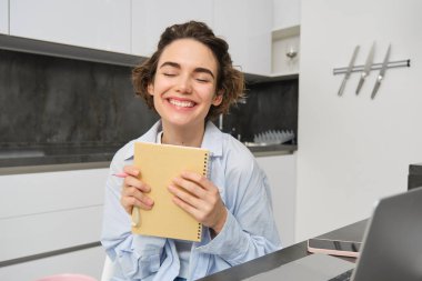 Gülümseyen esmer Womnan, günlüğünü tutuyor, planlayıcıyla yazıyor, dizüstü bilgisayarla evde çalışıyor, internetten çalışıyor ve not defterine notlar alıyor..