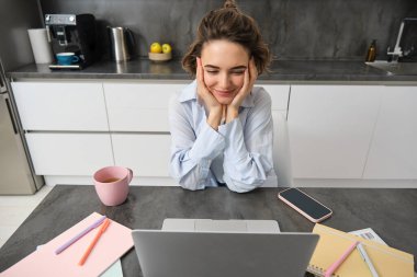 Evden çalışan bir iş kadınının en iyi görüntüsü. Kız uzaktan bilgisayarla çalışıyor, mutfakta oturuyor ve ekrana bakıyor, Webinar izliyor, online kurs..