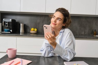 Teknoloji ve yaşam tarzı. Genç bir kadın evinde oturuyor, mutfağında akıllı telefon kullanıyor ve gülümsüyor..