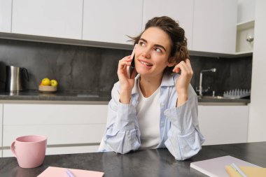 Gülümseyen genç bir kadın cep telefonuyla konuşur, evden birini arar, mutfakta oturur ve sohbet eder.