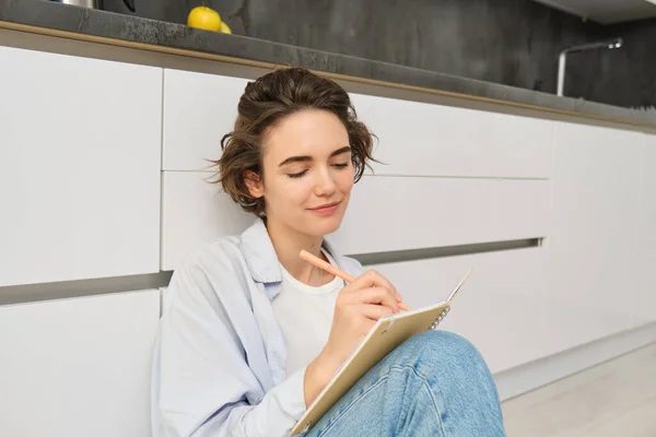Portrait of beautiful young woman writing in journal, adding notes in planner, sitting on floor and thinking, reads her diary, smiles with pleased face expression.