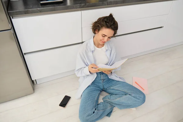 stock image Upper angle shot of young woman sitting on kitchen floor, revising for exam, reading her notes, doing homework at home.