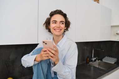 Gülümseyen esmer kız akıllı telefonuyla evde oturuyor, cep telefonu uygulamasından sipariş veriyor, online alışveriş uygulaması kullanıyor..