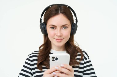 Esmer genç bir kadın resmi, gülümsüyor, kulaklıkla müzik dinliyor, cep telefonu uygulamasında videolar izliyor, akıllı telefonu tutuyor, beyaz arka planda duruyor..