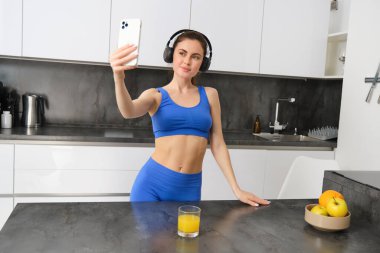 Tasasız sporcu kadın portresi, mutfakta akıllı telefonla dans etmek, kulaklıkla müzik dinlemek, antrenmandan sonra enerji dolu ve heyecanlı hissetmek..