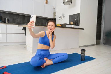 Genç gülümseyen bir kadın, fitess kız evden antrenman yapıyor, yoga minderinde akıllı telefonuyla oturuyor, selfie çekiyor, spor videolarını içeride kaydediyor..