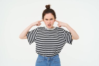 Sinirli, rahatsız bir kadın görüntüsü tıkanmış, parmaklarıyla kulaklarını kapatmış, gürültüden rahatsız olmuş, beyaz arka planda duruyor..