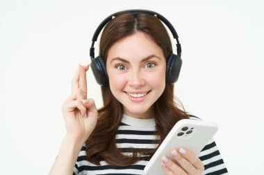 Parmaklarını çapraz tutan, dilek tutan, bir şey uman, akıllı telefon tutan, kablosuz kulaklık takan, beyaz arka plan.