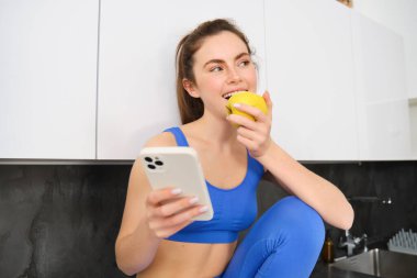 Sağlıklı, sportif genç bir kadının portresi. Akıllı telefonuyla mutfakta oturuyor, elma yiyor, meyve yiyor..