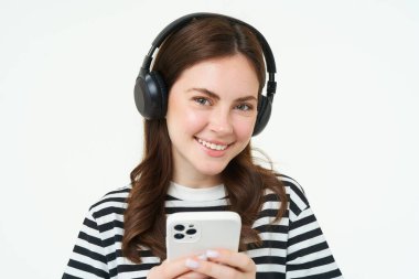 Esmer genç bir kadın resmi, gülümsüyor, kulaklıkla müzik dinliyor, cep telefonu uygulamasında videolar izliyor, akıllı telefonu tutuyor, beyaz arka planda duruyor..