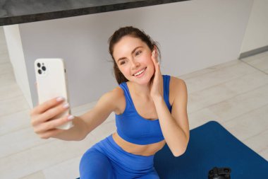 Güzel, atletik bir kız görüntüsü evde yoga minderinde oturuyor, akıllı telefonuyla selfie çekiyor ve egzersiz yapıyor, spor yapıyor..