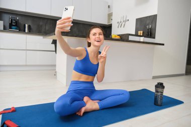 Aktif genç bir kadın, vlogger spor yapıyor, spor eğitimini evden kaydediyor, akıllı telefon kamerasıyla, evinde selfie için poz veriyor, mavi tayt ve spor sutyeniyle kauçuk yoga minderinde oturuyor..