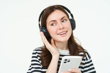 Gülümseyen, kulaklıklı güzel bayan öğrenci, cep telefonu tutuyor, gülümsüyor ve müzik dinliyor, akıllı telefon kullanıyor, beyaz arka planda duruyor..