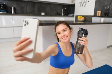 Refah ve spor. Güzel genç bir kadın akıllı telefondan selfie çekiyor, evden fitness eğitimi alıyor, su içiyor, susuz kalmıyor ve egzersiz yapıyor..