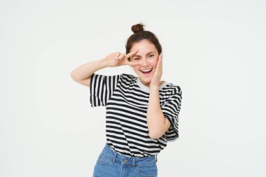 Güzel Avrupalı kadın fotoğraf için poz veriyor, barışı tutuyor, kawai el hareketi, kameraya gülümsüyor, beyaz arka planda duruyor..