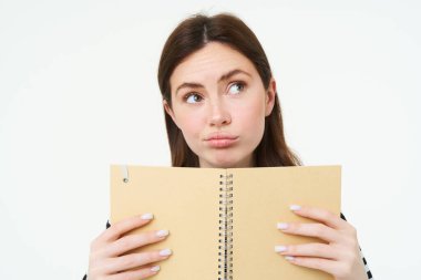 Elinde defter tutan düşünceli bir kızın portresi. Düşünceli bir yüzü olan genç bir kadın, haftalık planlayıcısını tutuyor, programını kontrol ediyor, not defterine notlar yazıyor, beyaz arka planda duruyor..