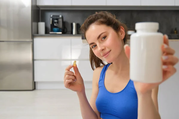 Close up portrait of young woman, fitness instructor showing bottle of vitamins, taking buds supplementary, dietary pill, sitting at home, doing workout exercises.