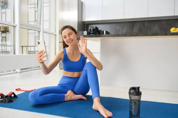 Portrait of young styling fitness girl doing workout from home, taking selfie and video for social media, gym instructor records her training session indoors.