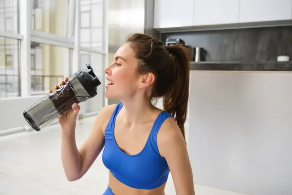 Portrait of sportswoman drinking water during workout session in living room, does her fitness training exercises at home.
